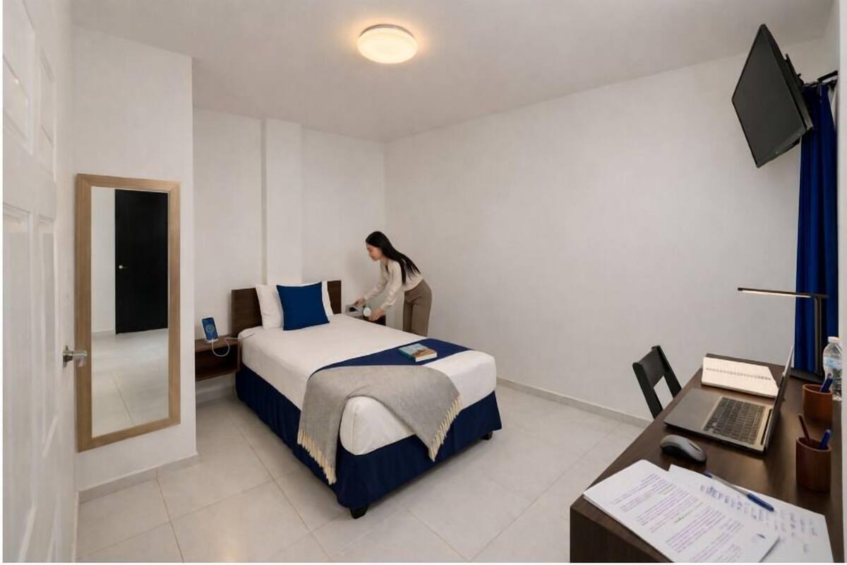 A hotel room with a bed, a desk with a laptop, a chair, a flat-screen TV, and a mirror.