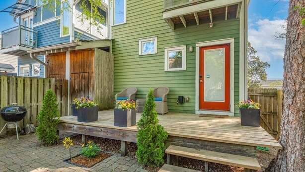 Une maison à deux étages, avec une façade verte, une porte d’entrée rouge et une terrasse en bois équipée de meubles pour se détendre à l’extérieur.