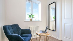 Una habitación con un sillón azul, una mesa redonda con una computadora portátil, una ventana con una planta y un espejo grande.