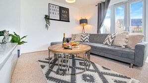 Una sala de estar moderna con un sofá gris, una mesa redonda con comida y bebidas, una lámpara y un ventanal con cortinas.