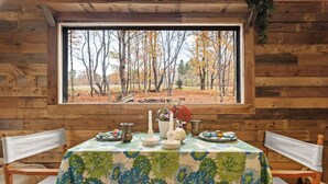 A rustic cabin interior with a dining table set for two, a window with a view of autumn trees, and wooden walls. - Off-Grid Tiny Cabin with Cozy Vintage Charm (Bovina)