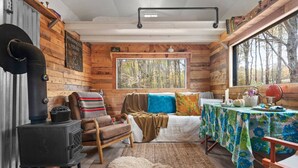 A cabin interior with a wood-burning stove, a sofa, a dining table with a floral tablecloth, and a large window overlooking a forest. - Off-Grid Tiny Cabin with Cozy Vintage Charm (Bovina)