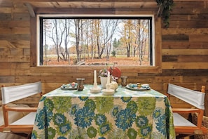 A wooden cabin with a table set for a meal, a window with a view of trees, and a tablecloth with a floral pattern.