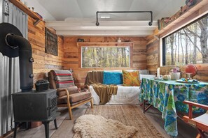 A cabin interior with a wood-burning stove, a cozy bed with colorful pillows, a dining table with a floral tablecloth, and a large window offering a view of trees.