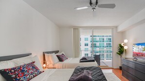 A hotel room with two beds, a ceiling fan, a large window with a city view, and a flat-screen TV on a dresser. - Stylish 1BR | Free Parking | Near Kaseya Center (Miami)