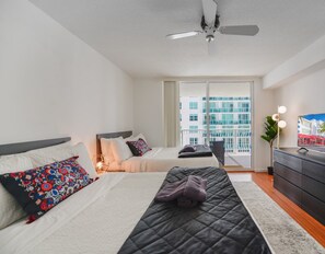 Une chambre d’hôtel moderne avec deux lits, un ventilateur de plafond, une grande fenêtre donnant sur la ville et un téléviseur à écran plat posé sur une commode.