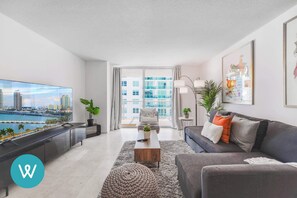 A modern living room with a large TV, a sofa, a coffee table, and a view of a cityscape through a large window.
