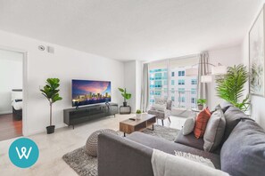 A modern living room with a large window, a flat-screen TV, a sofa, and a coffee table. - Stylish 1BR | Free Parking | Near Kaseya Center (Miami)