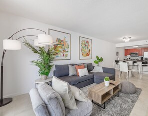 A modern living room with a grey sofa, a coffee table, and a floor lamp. There is a kitchen area with a dining table and chairs visible in the background.