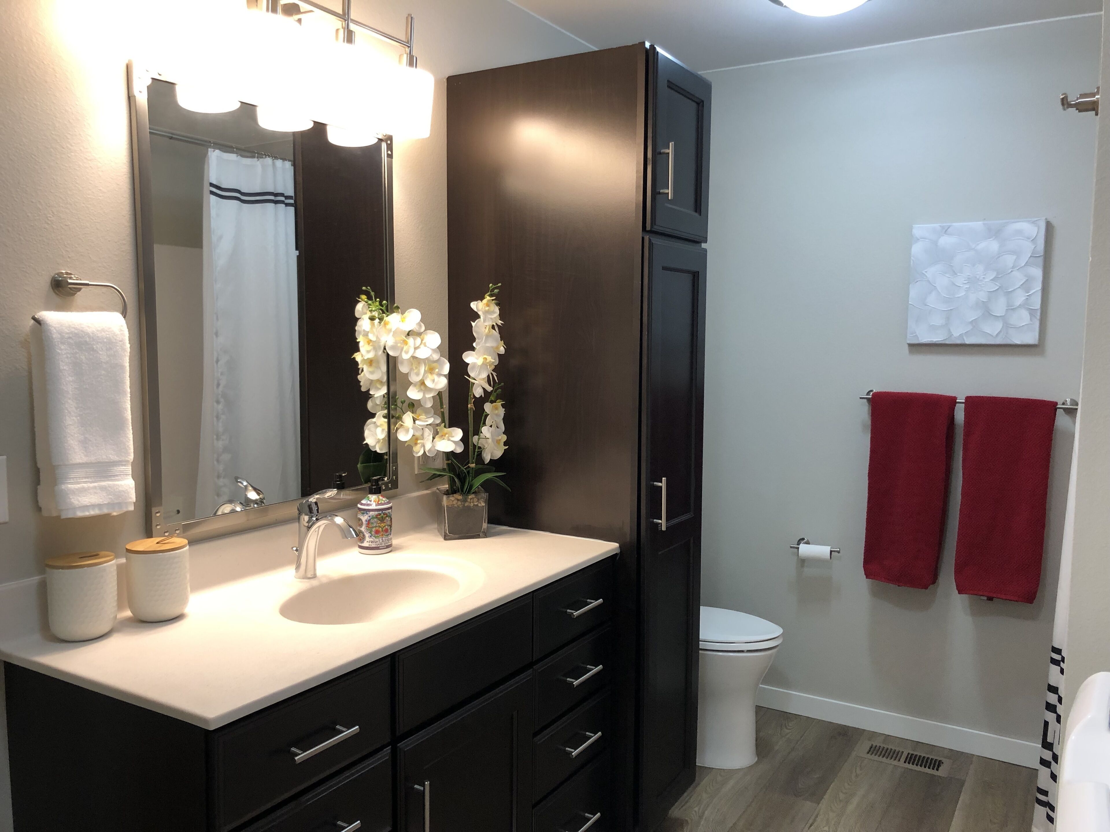 Une salle de bain moderne avec un meuble-lavabo en bois foncé, un lavabo blanc et un grand miroir. Deux serviettes blanches sont accrochées au mur et une serviette rouge est suspendue à un porte-serviettes.