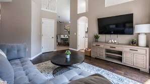 Una sala de estar con un sofá gris, una mesa de centro negra con una planta, un televisor sobre un mueble de madera y un área de comedor al fondo.
