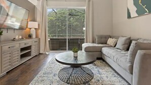 Una sala de estar moderna con una mesa de centro redonda de vidrio, un sofá seccional color beige, un televisor de pantalla plana y un ventanal con vistas a zonas verdes.