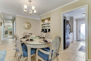 A dining area with a round glass table, wicker chairs with blue cushions, a chandelier, and a kitchen visible in the background.