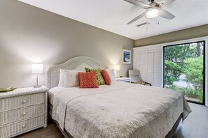 A bedroom with a bed, bedside tables, a ceiling fan, and a window with a view of greenery.