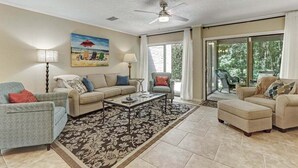 A spacious living room with a beige sofa, a grey armchair, and a coffee table. There is a painting of a beach scene on the wall, and a ceiling fan is visible. - Pet & EV-Friendly Fairway Oaks, Steps to Golf! (Fernandina Beach)