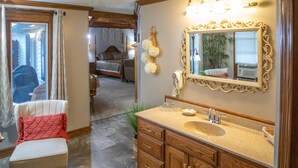 Une salle de bain avec un meuble-lavabo en bois, un grand miroir et un lit visible au fond.