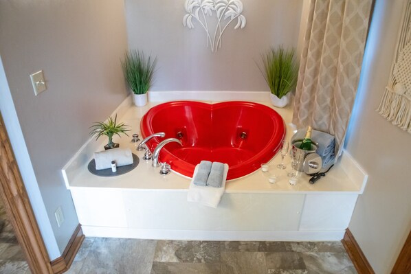 Une salle de bain avec une baignoire rouge en forme de cœur, un lavabo blanc, deux plantes en pot et un porte-serviettes.