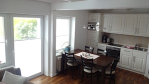 A modern kitchen with white cabinets, a dining table with chairs, and a view of greenery outside. - Cozy vacation home for up to 5 people in a quiet location in Greetsiel (Krummhörn)