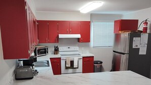 A compact kitchen with red cabinets, a white countertop, a microwave, a stove, and a refrigerator. There are two woven stools and a white chair at the counter.