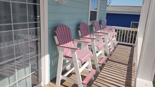 A covered porch with wooden deck and white railings, featuring several Adirondack chairs.
