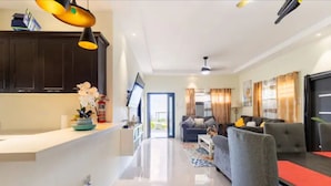 A modern living room with a grey sofa, a coffee table, and a TV. There is a kitchen area with dark cabinets and a white countertop.