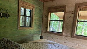 A wooden cabin room with a bed, two windows, and a view of trees.
