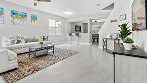 Un salon moderne avec un canapé blanc, une table basse et un tapis à motifs. On y trouve également un ventilateur de plafond, un téléviseur et un escalier menant à un autre niveau.