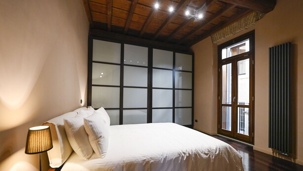 A bedroom with a bed, a nightstand, a lamp, a window with blinds, and a wooden ceiling. - Piazza Navona Mara’s apartment (Roma)