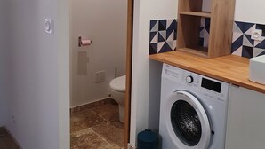 Ein kleiner Waschbereich mit Waschmaschine, einer kleinen Ablagefläche und einem WC, das durch eine offene Tür sichtbar ist.
