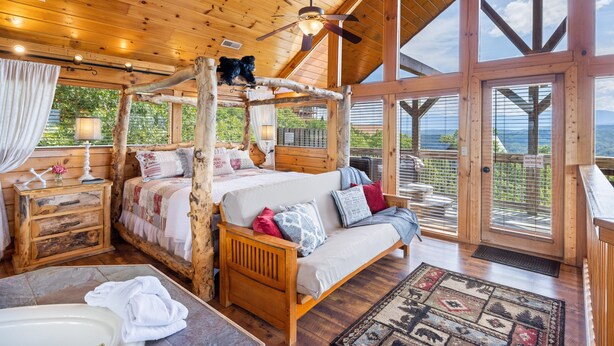 A wooden cabin with a four-poster bed, a wooden nightstand, a bathroom with a bathtub, and large windows offering a view of the outdoors. - High Mountain Mist - A Beautiful 4 Bedroom Cabin in Starr Crest Resort w/ Hot Tub and Pool Table! (Sevierville)