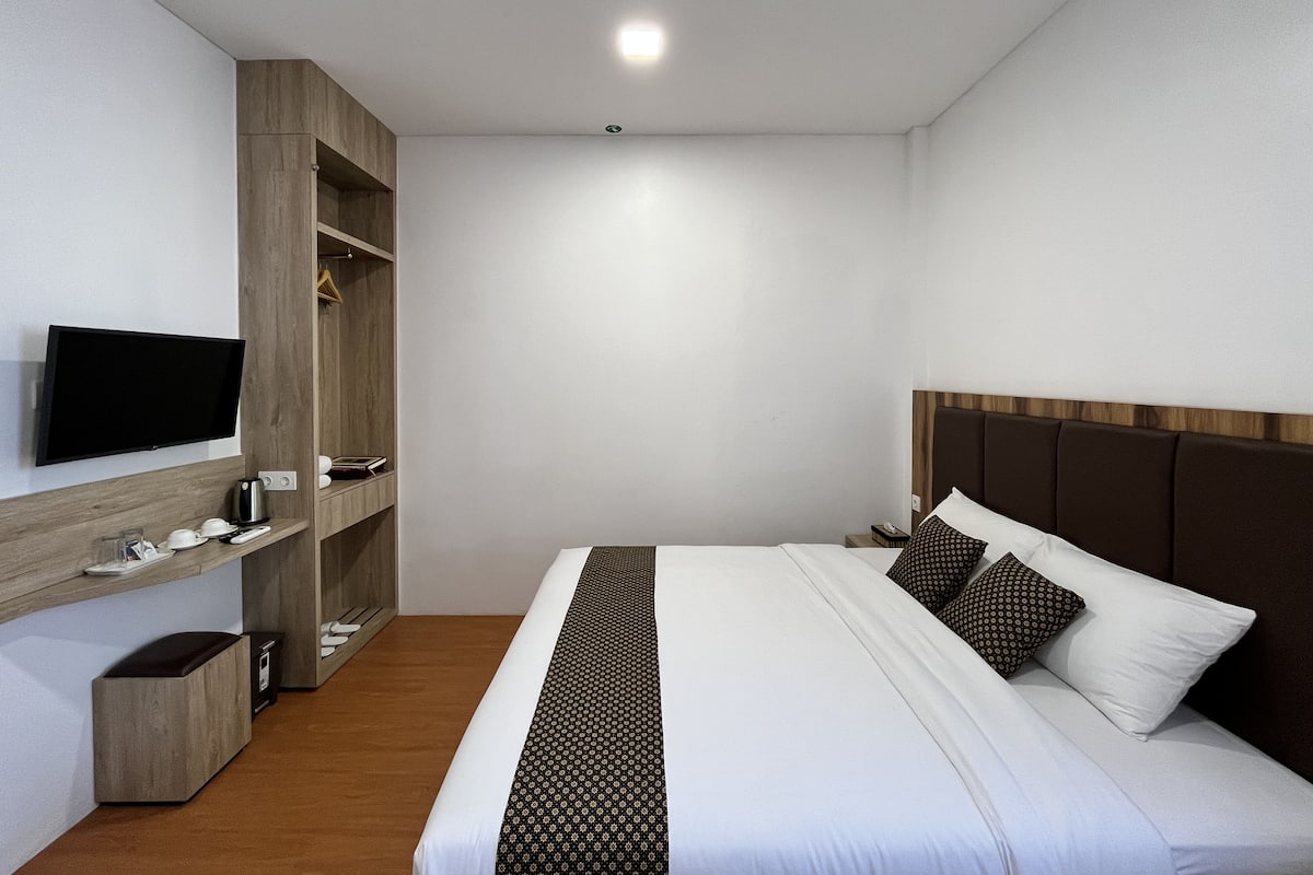 Quarto de hotel moderno com uma cama grande, televisão fixada na parede, guarda-roupa de madeira e mesa de cabeceira com abajur.