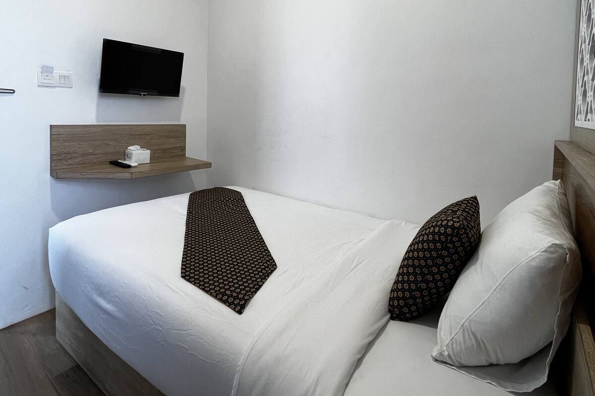Quarto de hotel moderno com uma cama de solteiro, televisão fixada na parede, mesa de cabeceira com um controle remoto e uma tomada de parede.