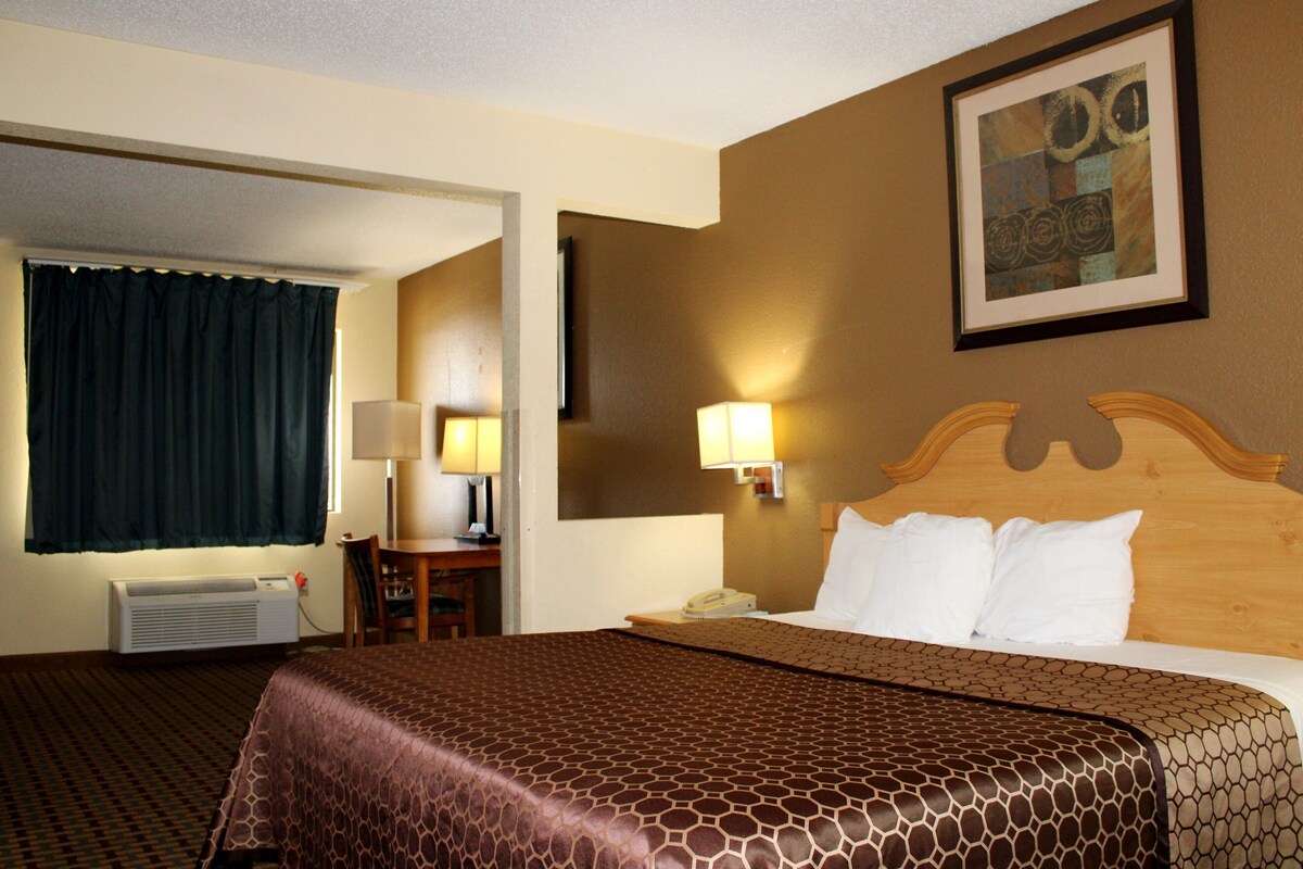 A hotel room with a bed, a desk, a chair, a lamp, a wall-mounted phone, and a framed picture on the wall.