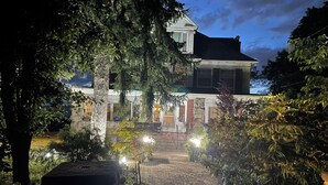 Un espacio exterior bien iluminado con un sendero de piedra que lleva a un edificio con porche. Se ven plantas en macetas y un perro pequeño.