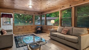 A cozy cabin interior with a wooden ceiling, large windows, a sofa, a glass coffee table, and a fireplace. - Charming Cozy Cabin by the Smoky Mtns. Game Room, Fireplace, Sunroom, Fire Pit (Sevierville)