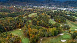 An aerial view of a golf course with multiple greens, fairways, and sand bunkers, surrounded by dense forest and a small town in the background. - Charming Cozy Cabin by the Smoky Mtns. Game Room, Fireplace, Sunroom, Fire Pit (Sevierville)