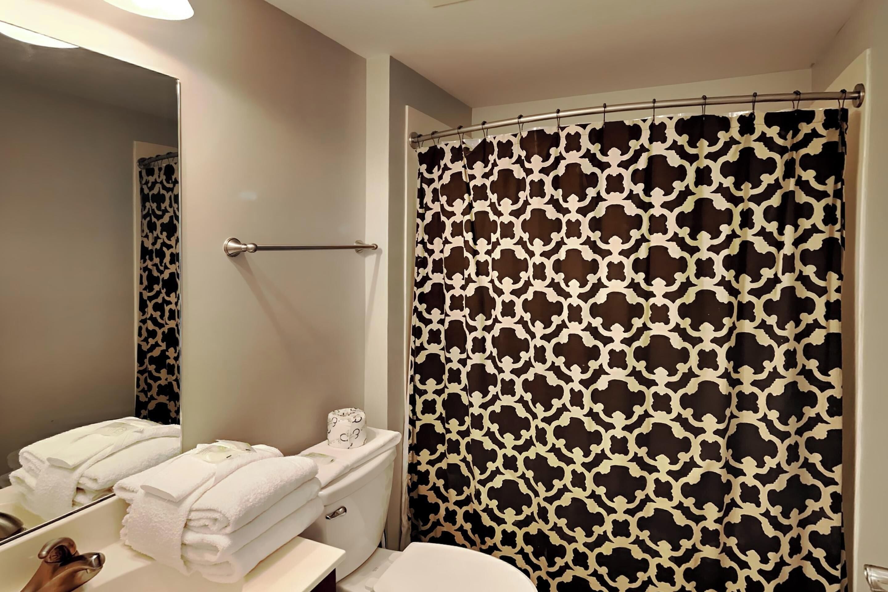 Un baño con inodoro, toallero con toallas dobladas, cortina de ducha con diseño geométrico y espejo que refleja el baño.