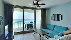 Una habitación con un sofá azul, una mesa de centro, un televisor y un ventilador de techo. Hay una puerta corrediza de vidrio que da a un balcón con vistas al océano.