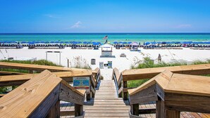 Un paseo marítimo de madera que conduce a una playa con hileras de sombrillas azules y sillas reclinables.