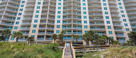 A high-rise hotel with multiple balconies, a sandy beach in the foreground, and a wooden staircase leading up to the entrance.