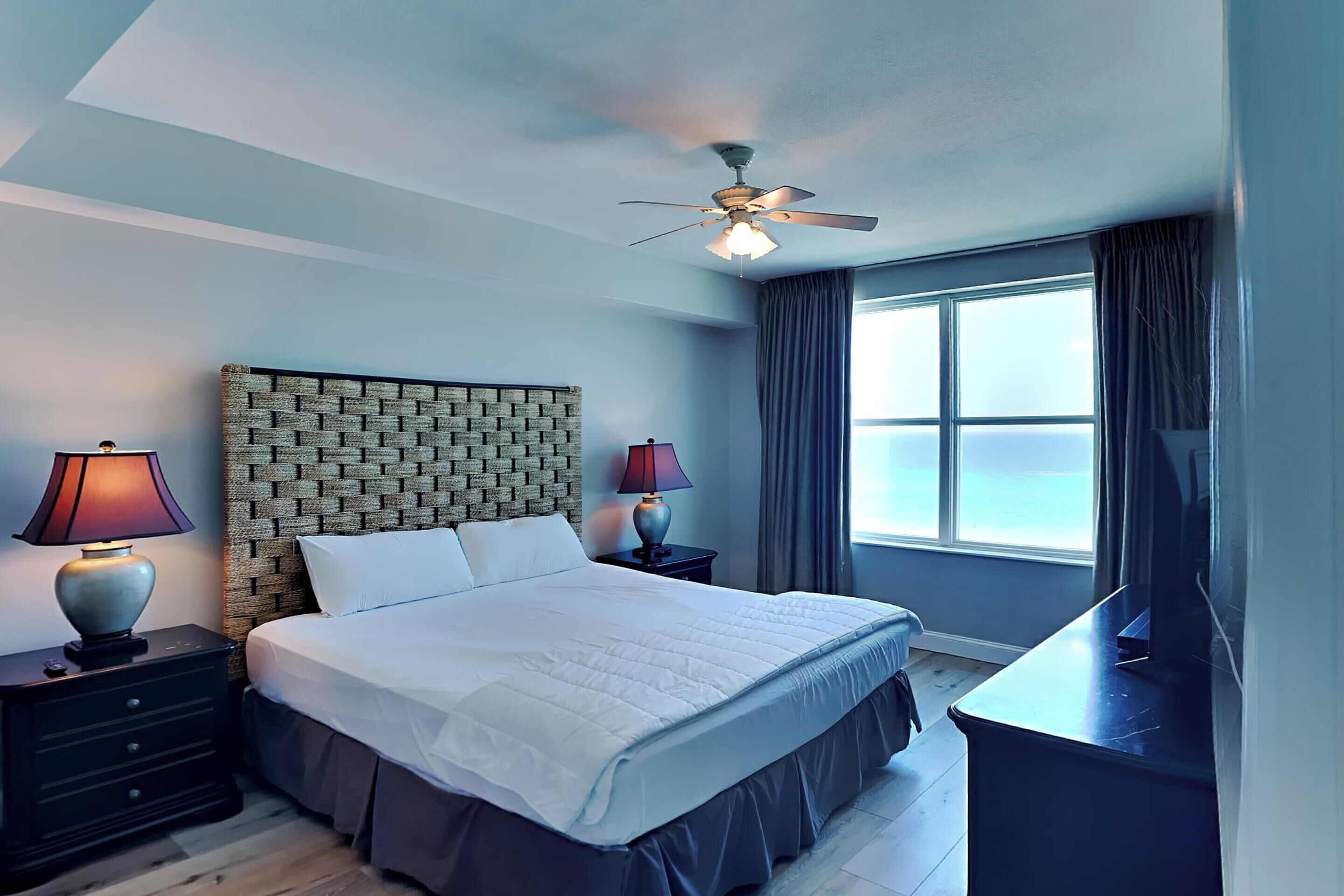 A hotel room with a brick-patterned headboard, a large bed with white linens, two bedside lamps, a ceiling fan, a large window with curtains, and a flat-screen TV on a dresser.