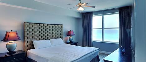 A hotel room with a brick-patterned headboard, a large bed with white linens, two bedside lamps, a ceiling fan, a large window with curtains, and a flat-screen TV on a dresser.