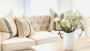 Un salon avec un canapé beige, des coussins blancs, un vase blanc orné de fleurs et une table basse en bois rustique.