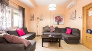 Una sala de estar con dos sofás grises, una mesa de centro y una lámpara de pie. Hay cuadros florales en las paredes y una puerta de madera con un letrero.
