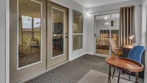 Una habitación de hotel moderna con una puerta de cristal corredera que da a un balcón, una pequeña zona de estar con una mesa y un ventanal grande con persianas.