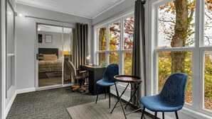 Habitación de hotel con cama, escritorio, dos sillas, una mesita y ventanas amplias con vistas a follaje otoñal.