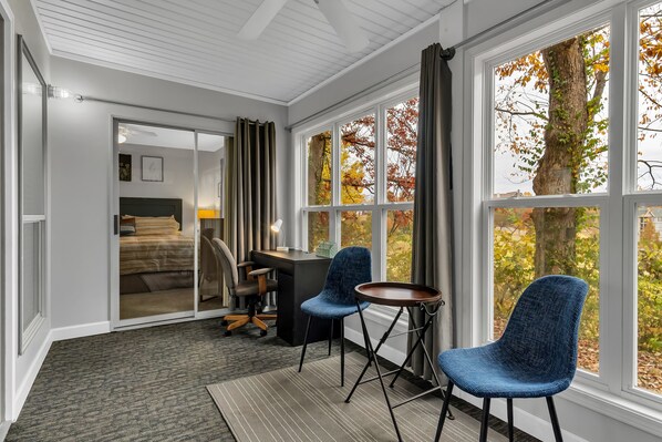 A hotel room with a bed, desk, two chairs, a small table, and large windows with autumn foliage views.