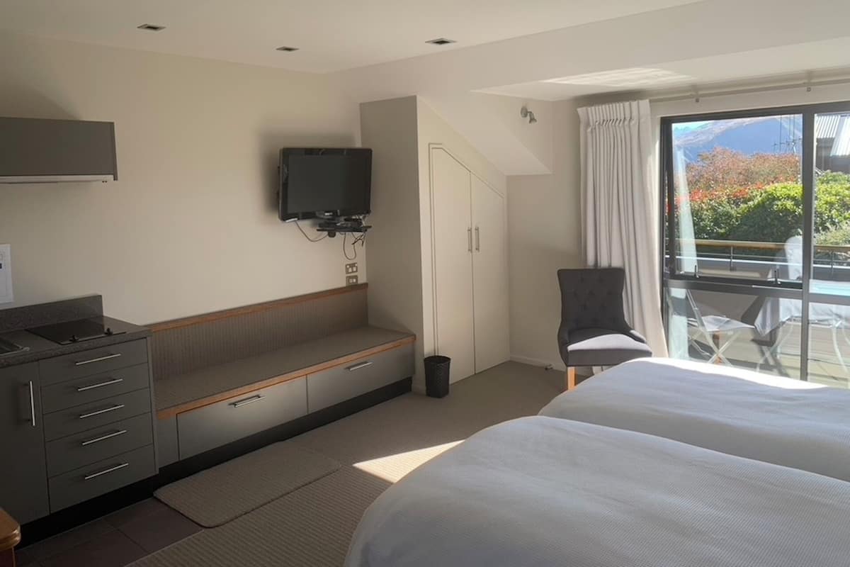 A hotel room with a bed, a TV mounted on the wall, a desk with drawers, a chair, and a window with a view of greenery.