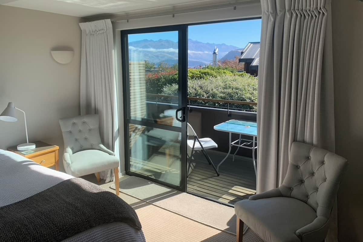 A hotel room with a bed, two chairs, a table, and a balcony with a view of mountains.
