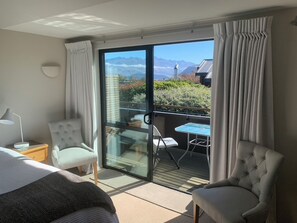 A hotel room with a bed, two chairs, a table, and a balcony with a view of mountains. - AspiringLofts (Wanaka)